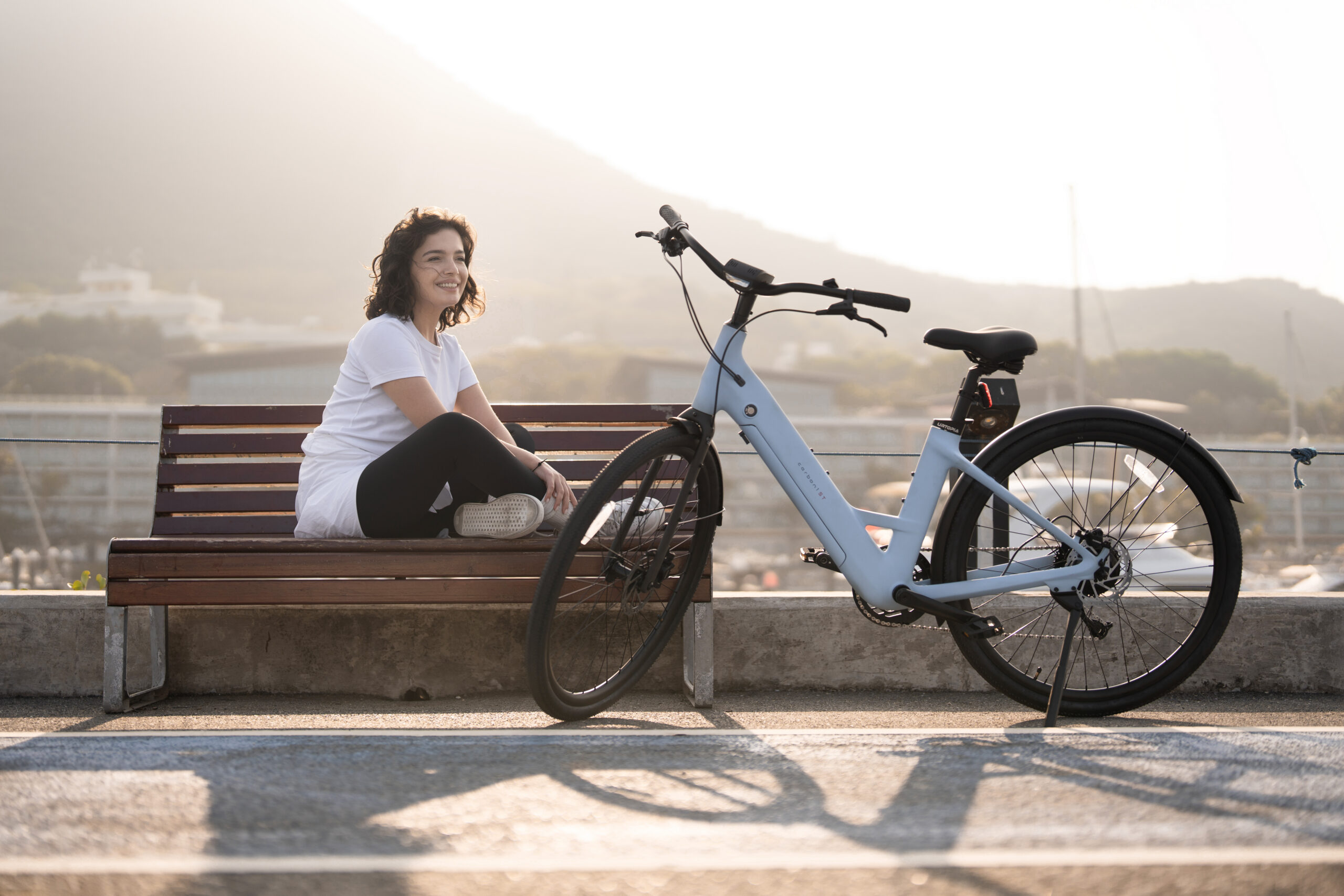 Woman sitting on a part bench next to a light blue Urtopia Carbon 1ST electric bike